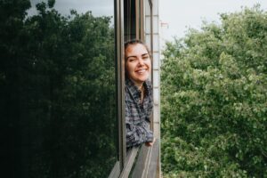 Усмихната жена гледа през прозорец на офис към зеленина